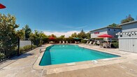 Located at 4934 San Juan Ave in Fair Oaks, California, this outdoor pool area offers a well-maintained communal retreat designed for relaxation and leisure. The centerpiece is a generously sized swimming pool with clear turquoise water, accessible via built-in entry steps and handrails. The pool features clearly marked depth indicators and posted safety signage, reflecting attentive facility management.

Surrounding the pool is a spacious concrete deck that provides ample room for lounging and socializing. The patio is furnished with multiple lounge chairs and chaise lounges positioned along the pool's perimeter, offering plenty of space for sunbathing. Several outdoor dining sets, each shaded by vibrant red and green umbrellas, are distributed around the deck, creating comfortable areas for outdoor dining or casual gatherings.

The pool area is fully enclosed by a wrought-iron safety fence with gated access, ensuring a secure environment. Mature trees and lush hedging line the perimeter, providing natural shade and a sense of privacy from the surrounding property. A storage structure is also visible within the enclosure. The overall setting benefits from an abundance of natural sunlight under open California skies, making it an inviting outdoor amenity well-suited for warm Fair Oaks summers.