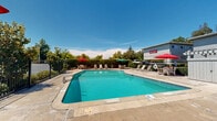 Located at 4934 San Juan Ave in Fair Oaks, California, this outdoor pool area offers a well-maintained communal retreat designed for relaxation and leisure. The centerpiece is a generously sized swimming pool with clear turquoise water, accessible via built-in entry steps and handrails. The pool features clearly marked depth indicators and posted safety signage, reflecting attentive facility management.

Surrounding the pool is a spacious concrete deck that provides ample room for lounging and socializing. The patio is furnished with multiple lounge chairs and chaise lounges positioned along the pool's perimeter, offering plenty of space for sunbathing. Several outdoor dining sets, each shaded by vibrant red and green umbrellas, are distributed around the deck, creating comfortable areas for outdoor dining or casual gatherings.

The pool area is fully enclosed by a wrought-iron safety fence with gated access, ensuring a secure environment. Mature trees and lush hedging line the perimeter, providing natural shade and a sense of privacy from the surrounding property. A storage structure is also visible within the enclosure. The overall setting benefits from an abundance of natural sunlight under open California skies, making it an inviting outdoor amenity well-suited for warm Fair Oaks summers.