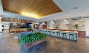 This single-floor amenity space makes a strong first impression with its blend of natural materials, contemporary design, and carefully curated details. The entry lobby features exposed brick walls, large windows that invite natural light, and a cozy seating arrangement of upholstered armchairs grouped around a round table — all anchored beneath a warm, illuminated coffered ceiling. A glass vestibule entry provides a clean transition from the outdoors.

Moving deeper into the space, a dramatic lounge area unfolds, anchored by a full-service bar with rich walnut cabinetry, white subway tile backsplash, and brass-accented open shelving adorned with potted greenery. Statement pendant lights with sculptural, wave-like shades hang overhead, adding an artistic focal point. A generously sized communal counter with bar stools runs alongside the bar, creating an inviting social hub.

Large planters overflowing with lush greenery are positioned throughout the open common area, adding warmth and a biophilic touch. Vertical wood slat screens provide elegant spatial definition between zones. Elevators clad in warm wood paneling offer access to upper floors, while a dedicated package locker room with herringbone-patterned tile flooring and rows of sleek smart lockers ensures practical convenience. The cohesive use of stone-look tile, warm wood tones, and layered lighting creates an elevated, welcoming atmosphere throughout.