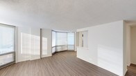This single-floor space showcases a clean, contemporary design with a neutral palette and an open, airy atmosphere. The standout architectural feature is a striking curved floor-to-ceiling window wall that floods the main living area with abundant natural light, casting warm, sweeping rays across the rich hardwood-style flooring. The smooth white walls contrast elegantly with the warm tones of the wood-look floors, creating a balanced and inviting interior.

The open layout of the main living area offers a generous sense of space and flexibility. A partial wall with a pass-through opening hints at a connected kitchen or service area beyond, providing a functional division while maintaining the open feel of the floor plan. The textured ceiling adds a subtle dimension to the otherwise streamlined design.

The curved window wall is particularly noteworthy, as it not only maximizes natural light but also lends the space a distinctive architectural character rarely seen in standard floor plans. Roller shades on the windows offer adjustable privacy and light control while preserving the clean, modern aesthetic. Overall, the space presents a bright, well-designed interior that combines thoughtful architectural details with a minimalist, versatile environment.