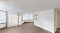 This single-floor space showcases a clean, contemporary design with a neutral palette and an open, airy atmosphere. The standout architectural feature is a striking curved floor-to-ceiling window wall that floods the main living area with abundant natural light, casting warm, sweeping rays across the rich hardwood-style flooring. The smooth white walls contrast elegantly with the warm tones of the wood-look floors, creating a balanced and inviting interior.

The open layout of the main living area offers a generous sense of space and flexibility. A partial wall with a pass-through opening hints at a connected kitchen or service area beyond, providing a functional division while maintaining the open feel of the floor plan. The textured ceiling adds a subtle dimension to the otherwise streamlined design.

The curved window wall is particularly noteworthy, as it not only maximizes natural light but also lends the space a distinctive architectural character rarely seen in standard floor plans. Roller shades on the windows offer adjustable privacy and light control while preserving the clean, modern aesthetic. Overall, the space presents a bright, well-designed interior that combines thoughtful architectural details with a minimalist, versatile environment.