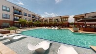 Located at 301 Silveron Blvd in Flower Mound, Texas, this outdoor amenity space offers a resort-inspired experience designed for relaxation and recreation. The centerpiece of the courtyard is a generously sized swimming pool with crystal-clear turquoise water, bordered by a spacious sun deck furnished with lounge chairs, chaise lounges, and shaded seating areas accented by bold blue market umbrellas and large decorative planters bursting with colorful flowers. The pool area includes a shallow wading zone with modern sculptural seating, ideal for lounging in the water on warm Texas days. A covered pergola structure anchors one end of the courtyard, providing a shaded gathering space with additional seating. Surrounding the pool, the deck transitions into a well-maintained green space featuring manicured lawns, ornamental trees, mulched garden beds, and winding pedestrian pathways that invite leisurely strolls through the landscaped grounds. The multi-story residential buildings that frame the courtyard feature a contemporary architectural design, blending neutral siding, brick accents, and large windows with private balconies that overlook the amenity area below. The entire outdoor space is thoughtfully designed to foster a sense of community while offering a tranquil, upscale retreat under open Texas skies.