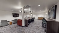 This contemporary communal lounge is thoughtfully designed to accommodate a variety of activities, from casual relaxation to collaborative gatherings. The open-plan layout is anchored by a richly patterned carpet in muted grey tones that adds texture and warmth underfoot throughout the entire space.

The lounge is divided into distinct functional zones. A central seating area features a bold red leather sofa paired with modern pod-style lounge chairs in soft grey, complemented by wooden stump accent tables and a grey upholstered ottoman. A wall-mounted flat-screen TV serves this area, making it well-suited for media viewing or casual socializing.

Along the perimeter, multiple bar-height counters offer additional seating with sleek dark stools, ideal for individual work or small group interaction. One counter area is highlighted by a large-scale abstract painting in deep blues and blacks, flanked by elegant oval wall sconces. A natural wood credenza provides additional storage and display surface nearby.

Recessed ceiling lighting is complemented by pendant fixtures and wall-mounted sconces, creating a layered and inviting ambiance. The crisp white walls keep the space feeling bright and open. The room also includes several high-top bistro tables with dark chairs, offering further flexible seating options throughout this well-appointed and versatile lounge.