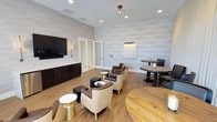 This single-floor lounge is designed with a contemporary aesthetic, combining functionality and comfort throughout its open layout. The walls are clad in light-toned, horizontal tile panels that lend a clean, textured backdrop to the entire space, complemented by warm hardwood flooring that runs throughout.

A focal point of the room is the media wall, where a large flat-screen television is mounted above dark, built-in cabinetry equipped with a compact refrigerator and flanked by wall sconces. This area creates an inviting entertainment zone suited for casual gatherings.

The seating arrangement is thoughtfully varied, featuring plush leather club chairs and accent ottomans grouped around gold-accented side tables, fostering intimate conversation areas. High-top bistro tables paired with sleek black chairs provide additional seating options suited for more upright, social interaction.

Floor-to-ceiling drapes line one side of the room, softening the space and framing large windows that introduce natural light and outdoor views. Recessed lighting and pendant fixtures overhead ensure consistent, well-distributed illumination throughout.

A set of frosted glass double doors offers access to adjacent rooms, maintaining an open yet defined sense of spatial flow. Overall, the lounge presents a polished, welcoming atmosphere ideal for social or professional use.