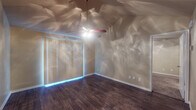 This single-floor space offers a thoughtfully laid-out interior with a warm, inviting ambiance throughout. The main living area immediately stands out with its vaulted ceiling, which lends a sense of openness and volume to the room. A ceiling fan with an integrated light fixture is centrally mounted, providing both air circulation and ambient illumination. The walls carry a subtle textured finish in a neutral beige tone, complemented by clean white baseboards that frame the rich, dark hardwood-style flooring — a consistent feature that flows through the living and adjoining areas. Large vertical blinds cover a sizable sliding door or window, allowing for flexible light control. A doorway from the main room leads to an additional carpeted space, adding variety in flooring and function. The galley-style kitchen is efficiently organized, featuring warm honey-toned wood cabinetry above and below the counters, a white electric range, a full-size refrigerator, a double-basin stainless steel sink, and a dishwasher. A window above the sink brings in natural light. The bathroom includes a full-size soaking tub with a shower, surrounded by clean white tile. The overall layout, as seen in the dollhouse view, presents a compact yet functional floor plan with well-defined living, kitchen, and private areas.
