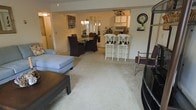 This single-floor space showcases an open-concept layout that connects the living room, dining area, and kitchen in one cohesive flow. The living room features plush, light-colored wall-to-wall carpeting and is furnished with a contemporary sectional sofa and a dark wood coffee table, creating a comfortable gathering area. The space transitions naturally into a formal dining area, where a glass-top table is surrounded by dark upholstered chairs, adding an elegant contrast to the soft, neutral palette of the walls and flooring. A white breakfast bar with matching bar stools serves as a stylish divider between the dining space and the kitchen, offering additional seating and a casual dining option. The kitchen, visible through a passthrough opening, features white cabinetry and appears well-lit by natural light from a nearby window. A chandelier over the dining area adds a decorative touch and warm ambiance. Decorative accents, including framed artwork and a wall clock, contribute to the overall polished, lived-in feel. The open layout and consistent flooring throughout create a sense of spaciousness and easy flow between functional areas.