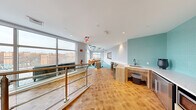 This stylish amenity lounge is thoughtfully designed to serve as a social and relaxation hub, elevated above the surrounding cityscape with sweeping urban views visible through expansive floor-to-ceiling windows. The space is divided into two distinct yet connected zones, unified by warm parquet hardwood flooring and a cohesive modern aesthetic.

The lounge area is an architectural standout, anchored by a striking coffered ceiling painted in teal and white, adding dramatic visual depth to the room. A plush, oversized orange sectional sofa paired with a sculptural coffee table and accent seating creates a vibrant, inviting gathering space. Aqua-toned area rugs complement the teal ceiling accents, while large windows on multiple walls flood the room with natural light and offer panoramic views of the surrounding urban skyline.

Transitioning toward the entry, a functional kitchenette features a built-in sink with a matte black faucet set against a bold teal vertical tile accent wall, alongside a compact counter with under-counter appliances including a refrigerator and microwave. A sleek glass-and-steel railing system defines the staircase landing, adding an open, airy quality to the space. Access to an outdoor terrace is visible through the lounge's glazed walls, extending the usable area beyond the interior and enhancing the overall sense of openness and light.