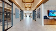 This elegantly designed single-floor community space makes a striking first impression with its expansive entry corridor, defined by soaring double-height ceilings, natural wood beam accents, and stacked stone columns. Large-format light tile flooring runs throughout, lending continuity and a clean, polished aesthetic. Black-framed glass doors line the corridor, while oversized curated artwork adorns the walls, setting an upscale tone from the moment of entry.

The main lounge area features a warm reclaimed wood plank ceiling with recessed lighting and exposed beams, paired with an eclectic mix of seating arrangements and a large area rug. A generous bank of windows along one wall floods the space with natural light and provides views of an outdoor patio area. An adjacent sitting nook offers a more intimate atmosphere, with a deep navy channel-tufted banquette, light gray accent chairs, and a trio of framed artworks illuminated by picture lights.

A refined secondary lounge showcases a bold slate-blue accent wall displaying four circular framed photographs, complemented by a coffered ceiling detail with warm wood inlay. The kitchenette area is equipped with rich walnut cabinetry, a large island with stone countertops, an integrated sink, and a wall-mounted display screen beneath a reclaimed wood ceiling. Throughout, stacked stone columns and floor-to-ceiling glass entry doors reinforce the sophisticated, modern-rustic character of the space.