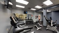 This fitness center is a comprehensive, single-floor facility designed to support a wide range of workout routines. The space features a clean, functional aesthetic with grey-painted walls, a drop ceiling with flush-panel fluorescent lighting, and durable rubber flooring throughout — a practical combination that keeps the environment bright and easy to maintain.

The cardio zone is anchored by a row of True-brand treadmills, each equipped with individual digital consoles, positioned along one wall beneath a wall-mounted television for entertainment during workouts. A wall-mounted speed bag adds a dynamic boxing element to this area.

The free weights section includes a fully stocked Icarian dumbbell rack with a wide range of weights, accompanied by adjustable weight benches for strength training. Large wall mirrors span this zone, enhancing the sense of space and allowing users to monitor their form.

The strength and cycling area houses True recumbent and upright stationary bikes alongside additional weight benches and cable machines. An Everlast heavy punching bag on a freestanding stand rounds out the boxing corner, offering a high-intensity training option.

The facility is accessed through frosted glass double doors etched with "Fitness Center," giving the entrance a professional look. Safety signage, hand sanitizer dispensers, and posted guidelines are visible throughout, reflecting a commitment to user safety and cleanliness.