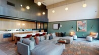 This thoughtfully designed open-plan space combines modern sophistication with warm, inviting materials across a generous single-floor layout. Dramatic double-height ceilings draw the eye upward, while a series of globe pendant lights provide warm, ambient illumination throughout. Floor-to-ceiling steel-framed windows flood the interior with natural light and offer views to an outdoor area, creating a seamless connection between indoors and out.

The main gathering area features a large, plush sectional sofa adorned with an array of decorative pillows, complemented by sleek leather lounge chairs with chrome frames and paired ottomans. A wall-mounted fireplace anchors one end of the room, adding both warmth and visual focus. The flooring transitions between large-format tiles and richly toned brick-pattern pavers, defining different zones within the open space.

A crisp white communal table with warm timber legs seats multiple guests alongside terracotta-toned upholstered chairs, ideal for group gatherings. Adjacent to this is a kitchenette finished with rich wood cabinetry, a striking veined quartz countertop, and integrated appliances. Deep teal-green accent walls provide a bold, cohesive backdrop throughout, enhanced by curated artwork and adjustable wall-mounted sconces. The overall ambiance is contemporary, warm, and highly functional.
