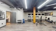 This single-floor fitness facility is designed to inspire and accommodate a full range of workout activities. The main exercise area features an open layout with durable wood-style flooring and an exposed ceiling with dark-painted structural beams and ductwork, lending the space an industrial aesthetic complemented by bright linear LED lighting overhead. A bold orange accent column and feature wall serve as visual focal points, reinforcing the facility's energetic branding.

The cardio section is lined with multiple treadmills and a cable machine, each positioned in front of wall-mounted flat-screen monitors for entertainment during workouts. A dedicated strength and free-weight area includes a multi-function cable/weight machine, adjustable benches, a dumbbell rack, and a selection of functional training accessories such as medicine balls, resistance equipment, and a freestanding punching bag. Two full-length mirrors are mounted along one wall, aiding form and adding depth to the space.

Motivational phrases are displayed prominently throughout, including "stronger than yesterday" and "you are only one workout away from a good mood," contributing to an uplifting atmosphere. A water fountain station is conveniently available within the workout area. The facility also includes a clean, fully accessible restroom with grab bars and wood-style flooring, ensuring comfort and compliance for all users.