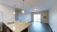 This single-floor space showcases a thoughtfully designed open-concept layout that seamlessly connects the kitchen and living areas. The kitchen is anchored by a generous island or breakfast bar finished with light granite-style countertops, offering both functional prep space and casual seating. Two stylish pendant lights hang above the counter, providing focused task lighting while adding a modern touch to the space.

The flooring throughout is a rich, dark hardwood-style material that contrasts elegantly with the neutral grey walls, creating a clean and contemporary aesthetic. The open living area benefits from natural light streaming in through a large sliding glass door fitted with vertical blinds, which leads to an outdoor space such as a balcony or patio.

A flush-mount ceiling fixture in the living area supplements the ambient lighting, while baseboard heating units along the walls indicate a practical and efficient heating system. A doorway visible in the background hints at additional rooms or storage within the unit. Overall, the space presents a modern, well-lit, and functional environment with a cohesive design palette.