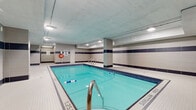 Located at 1551 Riverside Dr in Ottawa, Ontario, this indoor pool facility offers a spacious and well-maintained aquatic space spanning a generous footprint on a single level. The centerpiece is a rectangular swimming pool with vivid turquoise water, clearly marked depth indicators, and stainless steel entry railings for safe and convenient access. The pool is bordered by crisp white mosaic tile decking that extends around its perimeter, providing ample walkway space. The walls are finished in a two-tone large-format tile design, featuring neutral beige panels accented by a bold horizontal band of deep navy blue, lending the space a clean, modern aesthetic. Recessed and flush-mounted ceiling lights illuminate the area evenly, creating a bright and inviting atmosphere. Safety is well-addressed, with life rings mounted on the walls and prominent signage indicating pool depth and no-diving zones. A windowed observation or utility area is positioned along one wall, adding a practical element to the layout. The overall ambiance is functional yet refined, making this facility a well-equipped indoor swimming destination suitable for both recreational use and structured aquatic activities.