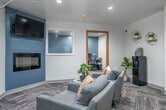 This well-appointed professional suite is thoughtfully designed to balance functionality with a welcoming ambiance. The space is divided into distinct zones, each serving a specific purpose while maintaining a cohesive contemporary aesthetic throughout.

The main lounge area serves as the heart of the suite, anchored by a sleek wall-mounted electric fireplace set against a steel-blue accent wall. A flat-screen TV is mounted above the fireplace, and a pair of plush grey armchairs arranged around a stylish side table — adorned with a bonsai plant — create an inviting atmosphere for guests or informal meetings.

Adjacent to the lounge, a dedicated workstation room is accessible through a warm wood-framed glass door. This focused workspace features a built-in desk accommodating multiple computer stations with ergonomic mesh chairs, making it ideal for productivity-driven tasks.

A secondary lounge area completes the suite, offering additional grey upholstered seating, a high-top table with industrial-style stools, and a coffee and refreshment station. Abstract wall art and a geometric wall clock add personality to the space.

Throughout, patterned herringbone carpet tiles, recessed lighting, and clean white walls contribute to a polished, professional environment that remains approachable and comfortable.