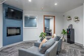 This well-appointed professional suite is thoughtfully designed to balance functionality with a welcoming ambiance. The space is divided into distinct zones, each serving a specific purpose while maintaining a cohesive contemporary aesthetic throughout.

The main lounge area serves as the heart of the suite, anchored by a sleek wall-mounted electric fireplace set against a steel-blue accent wall. A flat-screen TV is mounted above the fireplace, and a pair of plush grey armchairs arranged around a stylish side table — adorned with a bonsai plant — create an inviting atmosphere for guests or informal meetings.

Adjacent to the lounge, a dedicated workstation room is accessible through a warm wood-framed glass door. This focused workspace features a built-in desk accommodating multiple computer stations with ergonomic mesh chairs, making it ideal for productivity-driven tasks.

A secondary lounge area completes the suite, offering additional grey upholstered seating, a high-top table with industrial-style stools, and a coffee and refreshment station. Abstract wall art and a geometric wall clock add personality to the space.

Throughout, patterned herringbone carpet tiles, recessed lighting, and clean white walls contribute to a polished, professional environment that remains approachable and comfortable.