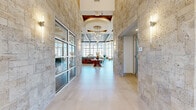 Located at 1600 LPGA Blvd in Daytona Beach, Florida, this sophisticated single-floor amenity and clubhouse space offers a generous, open layout designed to impress. The entrance corridor is defined by striking travertine stone wall cladding, large-format light tile flooring, and elegant wall sconces that set an upscale tone from the moment of entry. Floor-to-ceiling glass walls and expansive windows flood the interior with abundant natural light while framing views of the surrounding pool area and palm-lined grounds. The main gathering area features soaring double-height ceilings with a beautifully detailed coffered and wood-paneled ceiling, adorned by a dramatic chandelier that serves as a central focal point. A full-height stone fireplace anchors one end of the main lounge, which is furnished with multiple comfortable seating arrangements ideal for socializing or relaxing. An adjacent lounge area offers a more intimate setting with colorful artwork, a large-screen display, pendant lighting, and a communal dining table. Throughout the space, the interplay of natural stone columns, warm wood ceiling accents, and crisp white walls creates a refined yet welcoming Florida coastal aesthetic. The overall ambiance is bright, airy, and contemporary, making this a standout communal destination.