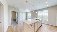 This 2-bedroom, 1-bathroom single-floor unit offers a thoughtfully designed open-concept layout that maximizes its compact footprint. The combined kitchen and living area serves as the heart of the home, anchored by a large central island with a white quartz countertop, an integrated sink, and seating space. The kitchen is outfitted with white shaker cabinetry, stainless steel appliances including a refrigerator, electric range, built-in microwave, and dishwasher. Three pendant lights hang above the island, adding a polished finishing touch. Light hardwood flooring runs throughout the main living areas, complemented by neutral beige walls that create a bright, airy atmosphere. Multiple windows, including a sliding glass door leading to an outdoor deck, invite generous natural light and outdoor views. The primary bedroom is a comfortable size, featuring plush carpet, a ceiling fan, and a walk-in closet with wire shelving. The second bedroom also offers hardwood-style flooring and a large window with outdoor views. The bathroom includes wood-look flooring, a vanity, and accessibility grab bars. A dedicated laundry area houses a stacked washer and dryer, adding everyday convenience to this efficiently designed home.