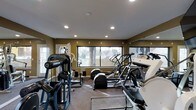 This fitness center offers a comprehensive workout environment designed to support a wide range of exercise routines. The space is outfitted with an impressive array of equipment, including elliptical machines, a treadmill, a cable weight machine, and additional strength training apparatus, providing options for both cardiovascular and resistance training.

Large windows line the walls, inviting abundant natural light into the room while offering views of the surrounding outdoor greenery. The warm, earth-toned walls create an energizing yet focused atmosphere, complemented by recessed ceiling lighting that ensures consistent illumination throughout. Full-length mirrors are positioned along one wall, enhancing the sense of openness and allowing users to monitor their form during workouts.

The flooring features durable carpet suited for a fitness environment, providing a degree of cushioning underfoot. The layout is thoughtfully arranged to maximize the available floor space, allowing for comfortable movement between equipment. The single-story design and well-maintained interior reflect a clean, functional space dedicated entirely to fitness and wellness.