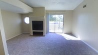 This single-floor space offers a welcoming and functional layout designed for comfortable living. The main living area is a standout, featuring plush wall-to-wall carpeting that adds warmth underfoot and a classic fireplace with a bold dark surround that serves as a natural focal point. A distinctive circular porthole-style window adds a unique architectural touch to the space, offering a charming contrast to the more traditional design elements throughout. Natural light floods the interior through a large sliding glass door, which opens to an outdoor view of mature trees and greenery, creating a seamless connection between the interior and the outdoors. The sliding door is dressed with vertical blinds, allowing for flexible light control. The textured ceiling adds subtle depth and character to the room. The neutral, light-toned walls provide a versatile backdrop that complements a wide range of furnishing styles. Overall, the space presents a clean, move-in-ready atmosphere with architectural details that add personality and charm to an otherwise classic and practical floor plan.