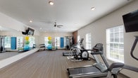 This single-story amenity space combines comfort and functionality across several well-designed areas. The main lounge serves as a central gathering point, featuring soaring ceilings, elegant chandelier lighting, and large glass-paneled entry doors that invite abundant natural light. Neutral-toned furnishings, including plush sofas and wingback chairs, are arranged to encourage both relaxed conversation and quiet retreat. A standout feature of the lounge is a floor-to-ceiling stone fireplace with a rustic wood mantle, adding warmth and a refined, lodge-inspired character to the space. French doors with black-trimmed grid-pane windows connect interior areas to the outdoors, maintaining a bright, airy atmosphere throughout. A casual dining area with a wooden table and chairs offers a functional spot for meetings or informal gatherings. The fitness center is well-equipped for a comprehensive workout, featuring treadmills, elliptical machines, cable strength equipment, and a dedicated stretching zone with stability balls and free weights. A ceiling fan, recessed lighting, and multiple windows keep the gym environment comfortable and well-lit. Collectively, the space presents a polished, community-oriented environment with a consistent palette of whites, neutrals, and natural stone textures.
