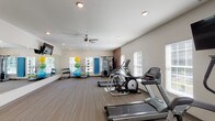 This single-story amenity space combines comfort and functionality across several well-designed areas. The main lounge serves as a central gathering point, featuring soaring ceilings, elegant chandelier lighting, and large glass-paneled entry doors that invite abundant natural light. Neutral-toned furnishings, including plush sofas and wingback chairs, are arranged to encourage both relaxed conversation and quiet retreat. A standout feature of the lounge is a floor-to-ceiling stone fireplace with a rustic wood mantle, adding warmth and a refined, lodge-inspired character to the space. French doors with black-trimmed grid-pane windows connect interior areas to the outdoors, maintaining a bright, airy atmosphere throughout. A casual dining area with a wooden table and chairs offers a functional spot for meetings or informal gatherings. The fitness center is well-equipped for a comprehensive workout, featuring treadmills, elliptical machines, cable strength equipment, and a dedicated stretching zone with stability balls and free weights. A ceiling fan, recessed lighting, and multiple windows keep the gym environment comfortable and well-lit. Collectively, the space presents a polished, community-oriented environment with a consistent palette of whites, neutrals, and natural stone textures.