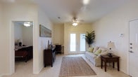 This well-appointed single-floor space offers a welcoming and functional layout designed for comfortable everyday living. The sitting room is a relaxed retreat, featuring light-toned carpet, a ceiling fan with integrated lighting, and a set of French doors that flood the room with natural light and provide access to an outdoor balcony. A neutral-toned sofa with accent pillows, a woven area rug, and a dark wood desk with storage create a versatile environment suitable for both lounging and working.

Transitioning into the interior, the dining area is anchored by a round bistro-style table surrounded by wrought-iron chairs, illuminated by an elegant chandelier overhead. Light-toned tile flooring extends throughout this portion of the space, lending a clean and airy aesthetic. The pale, neutral walls are accented by decorative artwork and carefully placed accent pieces, including a sculptural fleur-de-lis figurine atop a dark wood console.

Hallways connect to additional rooms, with glimpses of a bathroom visible in the background, suggesting a thoughtfully organized floor plan. Throughout the space, the consistent use of soft, warm tones, quality finishes, and carefully curated décor contribute to an inviting and cohesive ambiance.