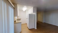 This single-floor space features a functional, open-concept layout that connects the kitchen and living areas seamlessly. The kitchen is outfitted with white upper and lower cabinetry, offering ample storage space, and includes a full-size white refrigerator and a stacked washer/dryer unit. A countertop with an undermount sink sits beneath a window, allowing natural light to filter in. The light-toned countertops complement the white cabinetry for a clean, cohesive look. The flooring throughout is warm hardwood-style laminate, lending continuity and visual warmth to the space. A textured ceiling with flush-mounted lighting provides even illumination. The kitchen flows naturally into an adjacent living area, which features the same flooring and leads to what appears to be a front entry door, enhancing the sense of connectivity within the layout. The overall color palette is neutral and bright, dominated by white walls and cabinetry, creating a clean and welcoming atmosphere throughout the space.