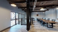 This single-floor commercial space masterfully blends industrial and modern design elements into a cohesive, functional work environment. The architecture is defined by exposed dark wood-plank ceilings with visible steel beams and overhead ductwork, creating a raw yet refined aesthetic throughout. Bold black steel structural columns and diagonal bracing serve as dramatic architectural features while reinforcing the industrial character of the space.

The main workspace is anchored by a large, solid wood conference table with herringbone detailing, surrounded by ergonomic task chairs — ideal for team meetings and collaborative sessions. A secondary standing-height black table provides an additional flexible work surface. Pendant lighting in an industrial barn-style hangs at varying heights, supplementing the recessed lighting to create a warm, inviting atmosphere.

Whitewashed vertical shiplap paneling and painted exposed brick walls add texture and contrast against the light, patterned hardwood flooring that runs throughout. A casual lounge area with contemporary upholstered chairs and a small coffee table offers a relaxed setting for informal gatherings, complemented by a wall-mounted display screen. Large floor-to-ceiling windows with black steel framing allow abundant natural light to flood the space, and a staircase with modern cable railings connects to the level below.