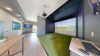 Located in Calgary, AB, this professionally designed golf simulator facility offers an immersive and technology-forward experience for golf enthusiasts of all skill levels. The space is thoughtfully divided into two functional zones that complement each other seamlessly.

The centerpiece of the facility is the dedicated simulator bay, which features lush artificial turf flooring and a designated hitting mat. A large-format projection screen spans the front wall, displaying detailed course visuals and real-time shot analytics, while an overhead projector and launch monitor system deliver accurate ball-tracking data. The bay is enclosed by sleek dark acoustic panel walls and a matching dark ceiling, creating a focused and immersive environment for play.

Adjacent to the simulator bay is a bright and welcoming lounge area finished with warm light wood flooring, neutral grey walls, and recessed lighting. This space is furnished with a high-top bar table and stools, ideal for spectators or for reviewing game data between sessions. A wall-mounted flat-screen TV, a large landscape art piece depicting a scenic aerial view, and a small refrigeration unit round out the amenities. A window at the far end of the lounge brings in natural light and views of the surrounding Calgary landscape, adding an airy, relaxed atmosphere to the overall space.