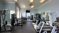 Located at 2600 Harvest Creek Pl in Cary, NC, this fitness center offers a well-equipped, single-floor workout facility designed to support a comprehensive exercise routine. The room is finished with soft blue walls, white crown molding, and a dark rubberized floor, creating a clean and functional athletic environment. Natural light streams in through multiple windows fitted with blinds, complemented by overhead flush-mount lighting and a ceiling fan that keeps the space comfortable during workouts. A wall-mounted flat-screen television provides entertainment while exercising. Full-length mirrors line one wall, visually expanding the space and allowing users to monitor their form. The equipment layout is thoughtfully organized, featuring a dedicated free weights section with a rack of dumbbells and a weight bench, as well as a strength training cable machine. The cardio section includes treadmills, ellipticals, and additional aerobic machines positioned to take advantage of the natural light from the windows. The combination of ample equipment variety, bright surroundings, and practical amenities makes this fitness center a convenient and fully functional space for residents looking to maintain an active lifestyle.