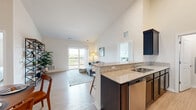 Located at 77 S Adams St in Oswego, Illinois, this well-appointed single-floor home offers three bedrooms, two bathrooms, a dedicated laundry area, and a thoughtfully designed layout. The open-concept kitchen and living area serves as the heart of the home, featuring striking vaulted ceilings that enhance the sense of space. The kitchen is equipped with dark cabinetry, granite countertops, stainless steel appliances — including a refrigerator, gas range, microwave, and dishwasher — and a breakfast bar with seating, ideal for casual dining. The living room flows naturally from the kitchen and is bathed in natural light from a large sliding glass door that opens onto a private balcony overlooking the surrounding landscape. Light wood-style flooring runs throughout the main living areas, adding warmth and continuity. The spacious primary bedroom features plush carpeting, vaulted ceilings, and generous natural light. Additional bedrooms are equally comfortable, each with carpet flooring and windows. The two bathrooms are clean and functional; one includes a glass-enclosed walk-in shower. A dedicated laundry area and closet provide practical storage solutions, completing this efficient and inviting home.