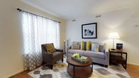 This single-story space at 8600 Glen Myrtle Ave offers a thoughtfully designed interior with a warm and welcoming ambiance. The living area serves as a central gathering space, showcasing a neutral color palette accented with cheerful yellow and gray tones that lend the room a modern, coordinated aesthetic.

Natural light flows generously into the living area through large windows dressed with elegant sheer patterned curtains, creating an airy and bright atmosphere. The space is furnished with a plush gray sofa adorned with a mix of decorative pillows in yellow, gray, and blue, complemented by a coordinating upholstered armchair with bold patterned cushioning. A round wooden coffee table anchors the seating arrangement atop a geometric patterned area rug, adding visual interest and warmth to the hardwood flooring beneath.

Wall décor includes a framed piece of artwork that adds a finishing touch to the space. Overhead, standard-height ceilings with visible HVAC vents ensure year-round comfort. The overall atmosphere is clean, contemporary, and move-in ready, reflecting a cohesive interior design sensibility throughout.