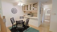 This single-floor space showcases a seamlessly integrated kitchen and dining area designed for functional, everyday living. The kitchen is appointed with light-toned wood cabinetry featuring glass-front upper cabinets, offering both display storage and an airy, open feel. A built-in cooktop, sink, and generous counter space are neatly arranged along the kitchen wall, while a peninsular breakfast bar creates a natural transition into the dining area. The dining space comfortably accommodates a round table paired with dark rattan chairs, anchored by a teal area rug that adds a pop of color against the warm-toned hardwood flooring that runs throughout. A ceiling fan with an integrated light fixture overhead provides both airflow and ambient lighting. Crown molding along the ceiling adds a refined architectural touch, while a hallway visible beyond the kitchen suggests additional rooms and living spaces further into the unit. Decorative wall accents, including a circular botanical piece and a wall-mounted metal sculpture, contribute character and warmth to the overall aesthetic. The space presents a clean, cohesive design with practical storage and a welcoming atmosphere.