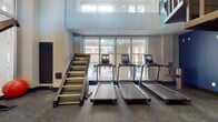 Located at 1312 S College St in Charlotte, NC, this two-floor fitness center is a well-equipped facility designed to support a comprehensive workout experience. The space is spread across two levels connected by a staircase, offering distinct zones for various types of exercise.

The lower level features an array of cardio equipment, including multiple treadmills and elliptical machines, positioned along floor-to-ceiling windows that flood the space with natural light and offer views of the surrounding neighborhood. Spin bikes and a cable machine add to the variety of equipment available. Durable rubber flooring runs throughout, providing both safety and comfort underfoot.

The upper level is dedicated to strength and functional training. Bold navy blue accent walls create a dynamic atmosphere, contrasting with lighter neutral tones. This area includes a dumbbell rack with a full range of free weights, medicine balls, TRX suspension trainers, resistance bands, and stability equipment. A wall-mounted flat-screen television and branded signage add to the polished finish of the space.

Large mirrors are strategically placed to enhance the sense of space and aid in form correction. The double-height ceilings in certain sections contribute to an open, airy ambiance. A chandelier visible through the interior glazing adds an unexpected touch of elegance to the facility's modern, functional design.
