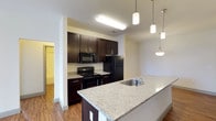 This well-appointed 3-bedroom, 2-bathroom single-floor residence offers a thoughtfully designed layout with modern finishes throughout. The heart of the home is an open-concept kitchen that seamlessly connects to the living areas, creating an inviting and functional space for everyday living and entertaining. The kitchen is highlighted by striking granite countertops, rich dark espresso cabinetry, and a large peninsula with a built-in sink — ideal for meal preparation or casual dining. Elegant pendant lighting hangs above the peninsula, adding a stylish focal point to the space. Full black appliances, including a refrigerator, range, and built-in microwave, provide a sleek, cohesive look. Warm hardwood-style flooring flows throughout the main living areas, complementing the contemporary color palette of neutral walls and dark wood tones. The open layout allows natural light to move freely through the space, enhancing the bright and airy ambiance. With three well-sized bedrooms and two full bathrooms, this residence offers ample space for comfortable living, making it a practical and attractive home for a variety of lifestyles.