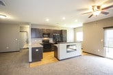 This single-floor space presents a cohesive, modern design anchored by a neutral gray color palette and consistent finishes throughout. The open-concept layout seamlessly connects the kitchen and living area, creating a generous, functional living space. The kitchen is well-appointed with dark espresso cabinetry, granite countertops, and a full suite of black appliances, including a side-by-side refrigerator with ice dispenser, electric range, built-in microwave, and dishwasher. Light hardwood flooring runs through the kitchen, complementing the dark cabinetry and providing a warm contrast. A peninsula with extended granite countertop serves as a natural divider between the kitchen and living area, while also housing a double-sided fireplace that adds both warmth and visual interest to the space. The living area features plush carpeting, recessed lighting, a ceiling fan, and a sliding glass door that opens to a private balcony, allowing for natural light and outdoor access. A separate room with ceiling fan and carpeted flooring offers additional flexible living space. The bathroom includes tile flooring, a granite-topped vanity with dark cabinetry, a walk-in shower, and neutral gray walls that maintain the home's consistent design aesthetic.