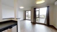 This single-floor space offers a clean, contemporary design with an open-concept layout that seamlessly connects the living area to the kitchen. The living space is bathed in natural light, courtesy of multiple large windows dressed with dark grommet curtains, providing both style and privacy. The windows offer views of the surrounding rooftops and green treetops, creating a pleasant urban-nature backdrop.

The flooring throughout is a warm, medium-toned hardwood-style material that adds continuity and a sense of warmth to the space. Flush-mount circular ceiling light fixtures provide even ambient illumination, complementing the abundant natural light.

The kitchen is partially separated from the living area by a low half-wall partition, topped with a dark stone countertop that mirrors the kitchen's countertop finish. This semi-open design maintains a sense of flow while defining the cooking zone. The kitchen cabinetry and appliances, visible along the left side, offer a functional and well-organized workspace.

The neutral, warm beige wall tones throughout create a versatile and welcoming atmosphere, suitable for a variety of interior styles. Overall, the space combines practicality with modern aesthetics, making it a well-lit and functional single-level residence.