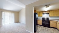 This single-floor residence offers a practical and comfortable layout designed for everyday living. The kitchen is outfitted with warm-toned natural wood cabinetry, light countertops, wood-look vinyl flooring, and black appliances including an electric range and a full-size refrigerator, complemented by an overhead range hood for ventilation. The kitchen opens toward the main living area, creating a connected and accessible flow throughout the space.

The living areas are finished with wall-to-wall carpeting in neutral tones, contributing to a warm and quiet atmosphere. Vaulted ceiling elements in portions of the unit add a sense of openness and architectural interest. A ceiling fan with light fixture is present, providing both air circulation and overhead lighting.

One of the rooms features a sliding glass door with vertical blinds, allowing natural light to filter in and providing access to an outdoor area. The entry door is solid and paneled, with secure hardware.

The bathroom includes a full-size tub and shower combination with clean white tile surround and chrome fixtures, offering a straightforward and functional design. Throughout the unit, neutral wall tones, clean lines, and consistent flooring choices create a cohesive and move-in ready environment.