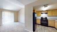 This single-floor residence offers a practical and comfortable layout designed for everyday living. The kitchen is outfitted with warm-toned natural wood cabinetry, light countertops, wood-look vinyl flooring, and black appliances including an electric range and a full-size refrigerator, complemented by an overhead range hood for ventilation. The kitchen opens toward the main living area, creating a connected and accessible flow throughout the space.

The living areas are finished with wall-to-wall carpeting in neutral tones, contributing to a warm and quiet atmosphere. Vaulted ceiling elements in portions of the unit add a sense of openness and architectural interest. A ceiling fan with light fixture is present, providing both air circulation and overhead lighting.

One of the rooms features a sliding glass door with vertical blinds, allowing natural light to filter in and providing access to an outdoor area. The entry door is solid and paneled, with secure hardware.

The bathroom includes a full-size tub and shower combination with clean white tile surround and chrome fixtures, offering a straightforward and functional design. Throughout the unit, neutral wall tones, clean lines, and consistent flooring choices create a cohesive and move-in ready environment.