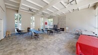 Located at 750 Mobil Ave in Camarillo, California, this versatile event and community hall offers a generously proportioned open floor plan ideal for gatherings, meetings, and multi-purpose use. The space is defined by its striking vaulted ceiling with exposed white-painted wooden beams, lending a sense of height and architectural character throughout. Multiple ceiling fans are suspended from the beam structure, ensuring comfortable airflow across the entire room. One of the most prominent architectural features is a floor-to-ceiling stone fireplace that anchors one end of the hall, providing a warm and inviting focal point. Large floor-to-ceiling windows line one wall, flooding the interior with abundant natural light and offering views of the mature trees and outdoor surroundings. The neutral-toned carpet tile flooring adds a practical yet polished finish to the space. The hall is equipped with a refrigerator and a flat-screen television, supporting a range of event and gathering needs. Multiple sets of white panel doors provide access points along the walls, enhancing circulation and connectivity. The open layout accommodates flexible furniture arrangements, currently shown with folding tables and chairs. This single-level space delivers a bright, airy, and functional environment well-suited for community events, classes, or social functions.