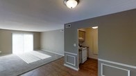 This single-floor unit offers a practical and comfortable layout designed to maximize usability. The open living area is bathed in natural light streaming through large windows with blinds, illuminating the combination of hardwood-style flooring and plush carpeted sections that define the space. Neutral gray and beige tones on the walls create a calm, cohesive ambiance throughout. A flush-mounted ceiling light provides additional illumination, complementing the bright natural light. The living area transitions seamlessly into a dedicated kitchen, separated by a distinctive gray accent wall with classic white wainscoting detail on a decorative column. The kitchen features warm wood-toned upper cabinetry, white electric range, and hardwood-style flooring, providing a functional and tidy cooking space. Ceiling-mounted vents are thoughtfully positioned for efficient climate control. White baseboards and trim throughout add a clean, finished look that ties each area together. The overall aesthetic balances simplicity and functionality, making this a well-appointed, move-in-ready space suited to everyday living.