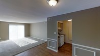 This single-floor unit offers a practical and comfortable layout designed to maximize usability. The open living area is bathed in natural light streaming through large windows with blinds, illuminating the combination of hardwood-style flooring and plush carpeted sections that define the space. Neutral gray and beige tones on the walls create a calm, cohesive ambiance throughout. A flush-mounted ceiling light provides additional illumination, complementing the bright natural light. The living area transitions seamlessly into a dedicated kitchen, separated by a distinctive gray accent wall with classic white wainscoting detail on a decorative column. The kitchen features warm wood-toned upper cabinetry, white electric range, and hardwood-style flooring, providing a functional and tidy cooking space. Ceiling-mounted vents are thoughtfully positioned for efficient climate control. White baseboards and trim throughout add a clean, finished look that ties each area together. The overall aesthetic balances simplicity and functionality, making this a well-appointed, move-in-ready space suited to everyday living.