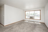 This single-floor unit offers a practical and straightforward layout across several rooms. The spacious living area is finished with neutral-toned carpet and white walls accented by warm oak wood trim, creating a clean and simple aesthetic. A large multi-pane window with a wall-mounted air conditioning unit allows generous natural light to fill the space while offering views of the surrounding exterior. Baseboard heating runs along the walls throughout, providing consistent climate control.

The additional carpeted rooms mirror the living area's design, each featuring wood-trimmed windows with blinds, neutral walls, and baseboard heaters. One room benefits from dual windows, enhancing its brightness and ventilation.

The kitchen is finished with tile flooring and features oak cabinetry providing ample upper and lower storage. A tile backsplash lines the cooking area, which includes a white electric coil-burner range with overhead ventilation. A double stainless steel sink is set into the countertop nearby. The overall layout is functional and efficiently organized. Throughout the unit, the consistent use of warm wood trim, neutral tones, and carpeted floors contributes to a cohesive and understated ambiance.