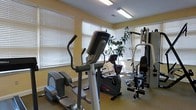 This single-floor fitness room is designed to support a well-rounded workout routine, offering a thoughtfully arranged selection of exercise equipment within a bright and welcoming environment. The space is outfitted with a Life Fitness elliptical trainer and a stationary upright bike, providing ample options for cardiovascular exercise. A multi-station cable and weight machine anchors one side of the room, enabling a variety of strength training movements. Multiple windows fitted with white horizontal blinds line the walls, allowing generous natural light to fill the space while offering privacy control. The warm yellow-toned walls and light-colored trim create an open and energizing atmosphere. Overhead fluorescent lighting supplements the natural light, ensuring the room remains well-lit throughout the day. A potted plant near the windows adds a touch of greenery, contributing to a refreshing ambiance. The carpeted flooring provides a comfortable, low-impact surface underfoot. Overall, this fitness room is a functional and inviting space that accommodates both cardio and strength training needs in a clean, organized setting.