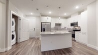 This single-floor residence in Bradenton, Florida, showcases a contemporary design with a clean, neutral color palette and wood-look flooring that flows consistently throughout the space. The kitchen serves as a central highlight, featuring a large island topped with speckled granite countertops, crisp white cabinetry with brushed chrome hardware, and a full suite of stainless steel appliances, including a French door refrigerator, electric range, and built-in microwave. Decorative pendant lights hang above the island, adding a modern touch to the open layout. A gray tile backsplash provides a subtle contrast to the white cabinetry, and recessed lighting ensures the space is well-illuminated. The bathroom continues the cohesive design aesthetic with an extended granite-topped vanity, white cabinetry, and a large mirror framed by overhead lighting. A walk-in shower with glass enclosure is also visible. The bedroom offers a bright, airy atmosphere, benefiting from natural light streaming through double windows fitted with blinds, while a ceiling fan contributes to the room's comfort. Overall, the space presents a modern, well-appointed interior with consistent finishes throughout.