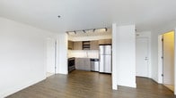 This single-floor space showcases a thoughtfully designed open-concept layout that seamlessly connects the living area to the kitchen. The kitchen is equipped with a full suite of stainless steel appliances, including a refrigerator, dishwasher, and range, complemented by warm, wood-toned cabinetry that extends to the upper walls for generous storage. Open shelving adds a modern touch, while a sleek countertop and undermount sink provide a functional workspace. Track lighting mounted to the ceiling illuminates the kitchen area with precision, enhancing both functionality and ambiance. The flooring throughout is a rich, dark hardwood-style material that grounds the space with warmth and continuity. Crisp white walls and ceilings create a bright, airy atmosphere that makes the space feel open and inviting. A structural column adds an architectural element that subtly defines the transition between the living and kitchen zones. The overall design aesthetic is clean, contemporary, and practical, making it an efficient and stylish living environment.