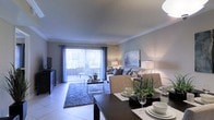 This single-floor residence features an open-concept layout that seamlessly connects the dining and living areas, creating a cohesive and functional living environment. The dining area is anchored by a dark-toned table set with place settings and a potted plant centerpiece, adding a touch of greenery to the space. Adjacent to the dining area, the living room is furnished with a neutral-toned sofa accented with decorative pillows, complemented by a textured area rug and a sleek glass coffee table. A piece of wall art adds a decorative focal point to the living space. The room benefits from abundant natural light streaming through large sliding glass doors fitted with vertical blinds and draped with elegant curtains, which lead to an outdoor area, enhancing the sense of openness. Light-toned tile flooring runs throughout, contributing to the bright and airy ambiance. Crown molding adds a refined architectural detail along the ceiling perimeter. The overall design palette of soft grays and warm neutrals creates a calm, inviting atmosphere throughout the space.