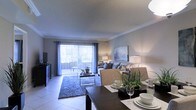 This single-floor residence features an open-concept layout that seamlessly connects the dining and living areas, creating a cohesive and functional living environment. The dining area is anchored by a dark-toned table set with place settings and a potted plant centerpiece, adding a touch of greenery to the space. Adjacent to the dining area, the living room is furnished with a neutral-toned sofa accented with decorative pillows, complemented by a textured area rug and a sleek glass coffee table. A piece of wall art adds a decorative focal point to the living space. The room benefits from abundant natural light streaming through large sliding glass doors fitted with vertical blinds and draped with elegant curtains, which lead to an outdoor area, enhancing the sense of openness. Light-toned tile flooring runs throughout, contributing to the bright and airy ambiance. Crown molding adds a refined architectural detail along the ceiling perimeter. The overall design palette of soft grays and warm neutrals creates a calm, inviting atmosphere throughout the space.