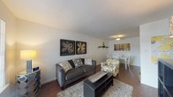 This single-floor residence offers a thoughtfully arranged layout that balances comfort and functionality. The open-concept living and dining area forms the heart of the home, featuring rich hardwood-style flooring that flows throughout the space. The living area is highlighted by neutral white walls that serve as a clean backdrop, complemented by decorative wall art and a cozy seating arrangement centered around a dark coffee table and a plush area rug. The adjoining dining space provides a dedicated area for meals, complete with overhead lighting and easy access to the kitchen. The kitchen is efficiently designed with warm wood cabinetry, dark granite-style countertops, and black appliances including a dishwasher and range, offering ample workspace for cooking. The two bedrooms are comfortably sized, each featuring hardwood-style flooring, patterned curtains, and generous natural light. One bedroom includes a window with an outdoor view, adding a sense of openness. The full bathroom is finished with classic white subway tile surrounding the shower-tub combination, complemented by brushed nickel fixtures. Throughout the space, clean lines and neutral tones create a cohesive and welcoming ambiance suited to everyday living.