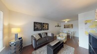 This single-floor residence offers a thoughtfully arranged layout that balances comfort and functionality. The open-concept living and dining area forms the heart of the home, featuring rich hardwood-style flooring that flows throughout the space. The living area is highlighted by neutral white walls that serve as a clean backdrop, complemented by decorative wall art and a cozy seating arrangement centered around a dark coffee table and a plush area rug. The adjoining dining space provides a dedicated area for meals, complete with overhead lighting and easy access to the kitchen. The kitchen is efficiently designed with warm wood cabinetry, dark granite-style countertops, and black appliances including a dishwasher and range, offering ample workspace for cooking. The two bedrooms are comfortably sized, each featuring hardwood-style flooring, patterned curtains, and generous natural light. One bedroom includes a window with an outdoor view, adding a sense of openness. The full bathroom is finished with classic white subway tile surrounding the shower-tub combination, complemented by brushed nickel fixtures. Throughout the space, clean lines and neutral tones create a cohesive and welcoming ambiance suited to everyday living.